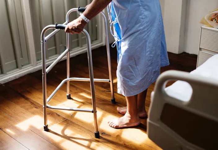 An older woman in a hospital gown standing barefoot on a wooden floor with both hands on a walking frame