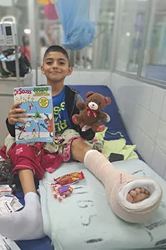 A boy sitting up on a hospital bed with a picture book and a teddy bear, one leg in a cast