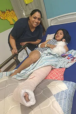 A girl resting in a hospital bed with a bandaged leg, a caregiver at her side