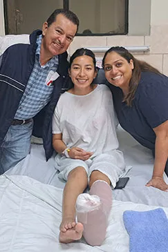 A young woman in a hospital gown sitting up in bed with two visitors beside her