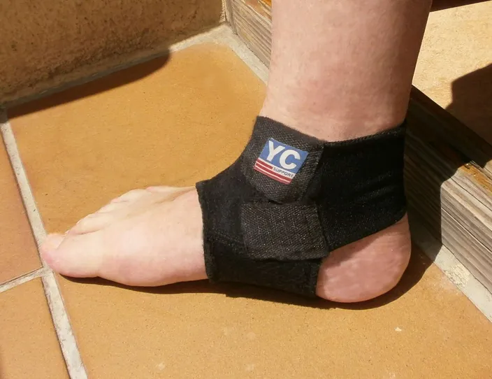A bare foot and ankle wearing a black fabric ankle brace fastened with a strap